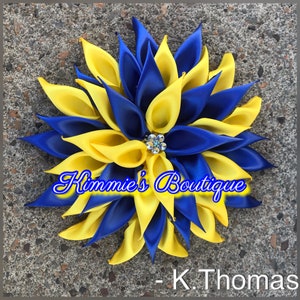 May include: A handmade, flower-shaped hair accessory with alternating yellow and blue satin petals. A sparkling rhinestone embellishment is at the center. The text "Kimmie's Boutique" and "- K. Thomas" are visible.
