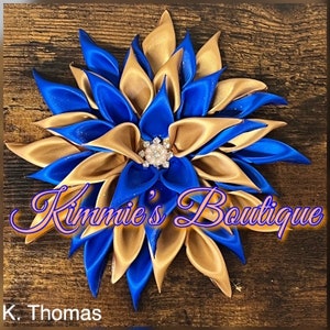 May include: A blue and gold satin flower with a pearl and rhinestone center. The flower is on a wooden background.  "Kimmie's Boutique" is written in pink script below the flower.  "K. Thomas" is written in black below the script.