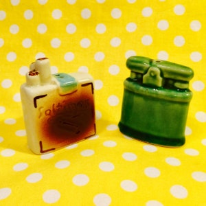 May include: A pair of ceramic salt and pepper shakers. One is a cream-colored rectangular shape with brown accents and the word "Saltines" on it. The other is a green, cylindrical shape with a lid. The background is yellow with white polka dots.