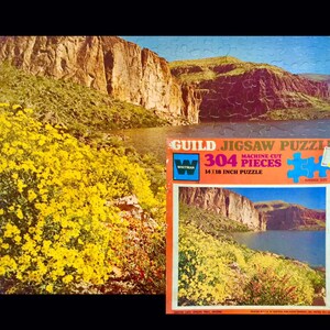 May include: A vintage Guild jigsaw puzzle featuring a scenic landscape of Canyon Lake, Apache Trail, Arizona. The puzzle, with 304 pieces, depicts a river scene with yellow flowers and rocky cliffs. The box measures 14 x 18 inches.