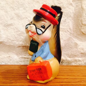 May include: A vintage ceramic squirrel figurine wearing a red hat, glasses, and a blue shirt. The squirrel holds a green book labeled "MAPS" and an orange suitcase with "BANK FOR VACATION FUND" printed on it. The figurine is on a wooden surface.