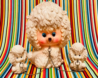 May include: A set of three vintage poodle figurines in off-white with blue eyes and pink cheeks, connected by a silver chain. The poodles have curly fur and are set against a colorful striped background.