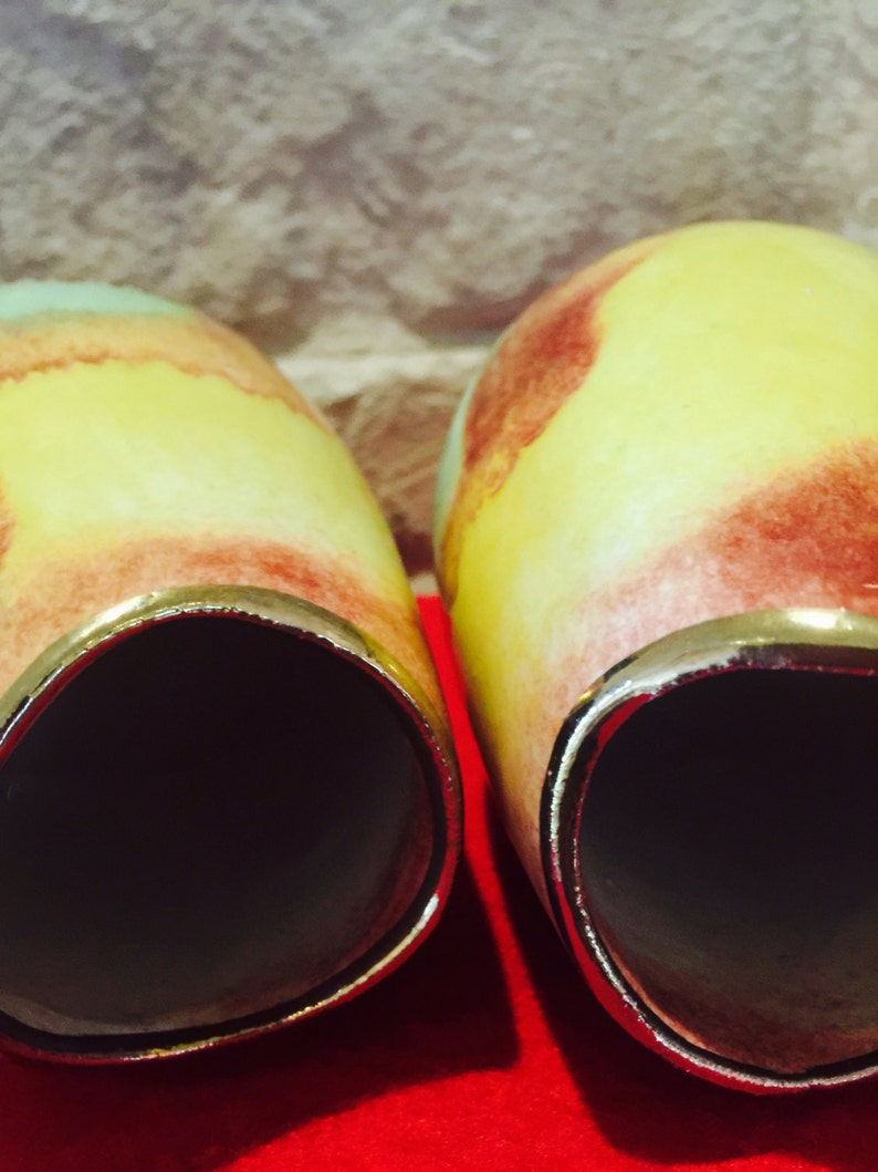 May include: Two ceramic vases with a yellow, red, and green color scheme. The vases have a gold rim and a dark interior. The vases are set against a neutral background and a red surface.
