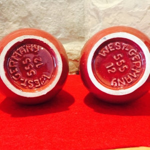 May include: Two round, red ceramic objects with white rims. The bottoms display the text "WEST-GERMANY 555 12" in a circular pattern. The objects sit on a red surface.