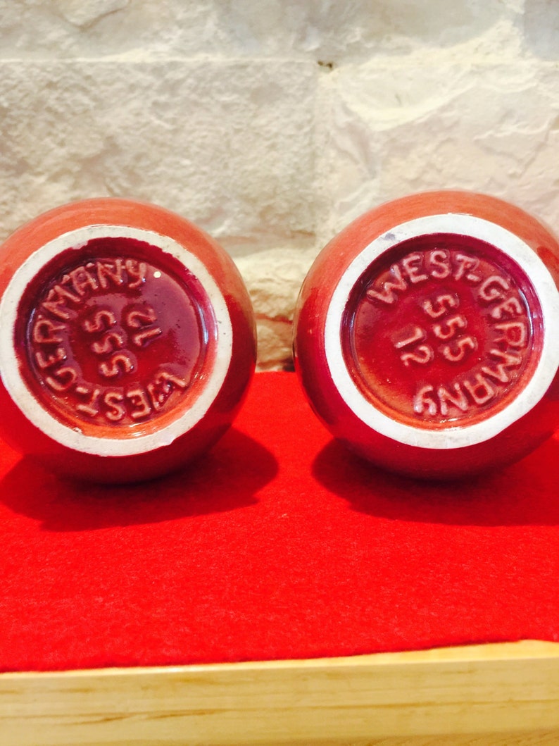 May include: Two round, red ceramic objects with white rims. The bottoms display the text "WEST-GERMANY 555 12" in a circular pattern. The objects sit on a red surface.