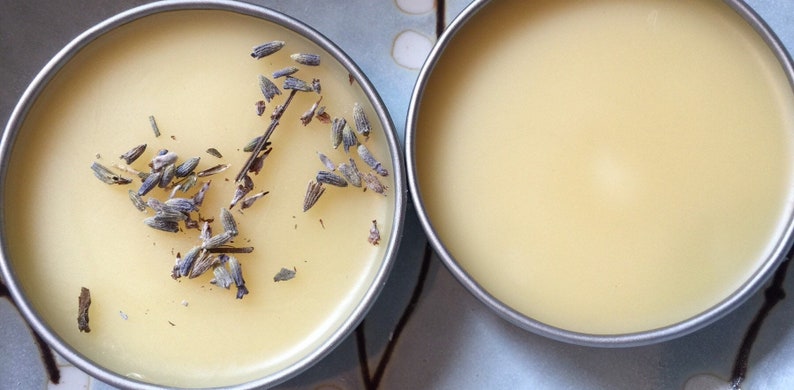K&ouml;nnte beinhalten: Zwei kleine runde Metalldosen gef&uuml;llt mit einem hellgelben Balsam. Eine Dose hat getrocknete Lavendelbl&uuml;ten auf dem Balsam.