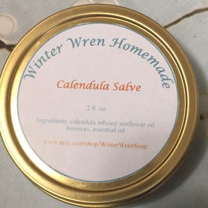 K&ouml;nnte beinhalten: Eine goldene Metalldose mit einem wei&szlig;en Etikett, auf dem steht: "Winter Wren Homemade Calendula Salve 2 fl oz. Ingredients: calendula infused sunflower oil, beeswax, essential oil www.etsy.com/shop/WinterWrenSoap"
