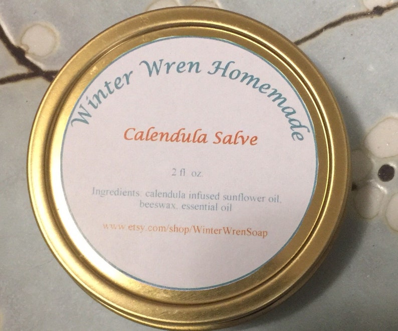 K&ouml;nnte beinhalten: Eine goldene Metalldose mit einem wei&szlig;en Etikett, auf dem steht: "Winter Wren Homemade Calendula Salve 2 fl oz. Ingredients: calendula infused sunflower oil, beeswax, essential oil www.etsy.com/shop/WinterWrenSoap"