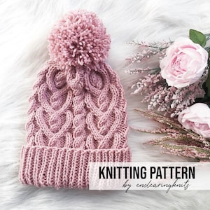 May include: A pink knitted hat with a cable knit pattern and a fluffy pom pom on top. The hat is on a white fluffy surface with pink flowers.