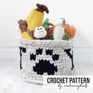Peut inclure: Un panier de rangement en crochet avec un motif de patte de chien en noir et blanc. Le panier est rempli de jouets en peluche, dont une banane, un singe et un jouet rouge et vert.