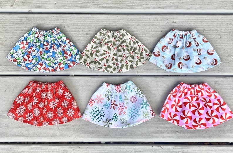 Puede incluir: Seis faldas de mu&ntilde;eca diferentes con tem&aacute;tica navide&ntilde;a. Las faldas est&aacute;n hechas de tela y tienen varios patrones, incluyendo bastones de caramelo, copos de nieve, acebo y Pap&aacute; Noel.