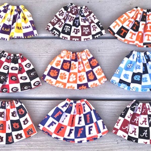 May include: Nine different fabric doll skirts with various college logos and colors. The skirts are checkered with white and a variety of colors, including red, blue, orange, purple, and black. The logos include LSU, Georgia, Clemson, Alabama, Florida, Tennessee, and Auburn.
