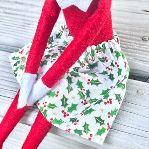 Puede incluir: Una mu&ntilde;eca de elfo roja con una camisa blanca de cuello y una falda verde y blanca con un patr&oacute;n de acebo. La falda tiene bayas rojas y detalles dorados.
