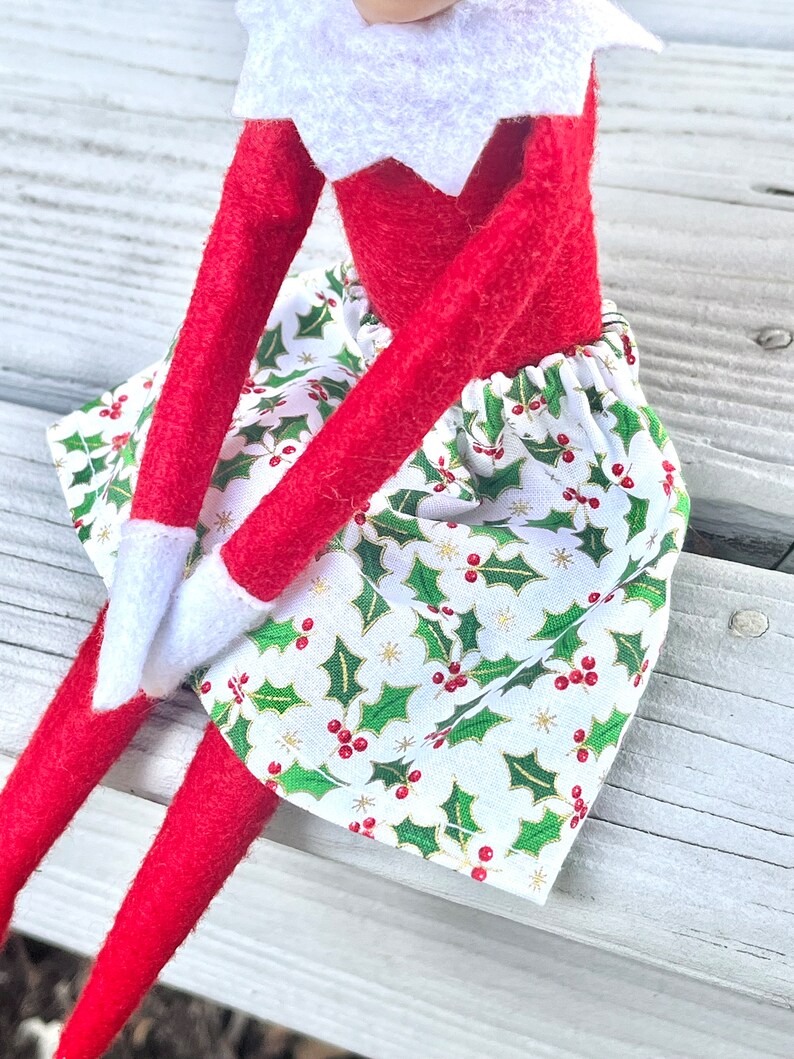 Puede incluir: Una mu&ntilde;eca de elfo roja con una camisa blanca de cuello y una falda verde y blanca con un patr&oacute;n de acebo. La falda tiene bayas rojas y detalles dorados.