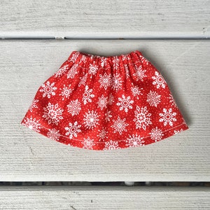 Puede incluir: Una falda roja con un estampado de copos de nieve blancos. La falda est&aacute; hecha de una tela suave y c&oacute;moda y tiene una cintura el&aacute;stica.