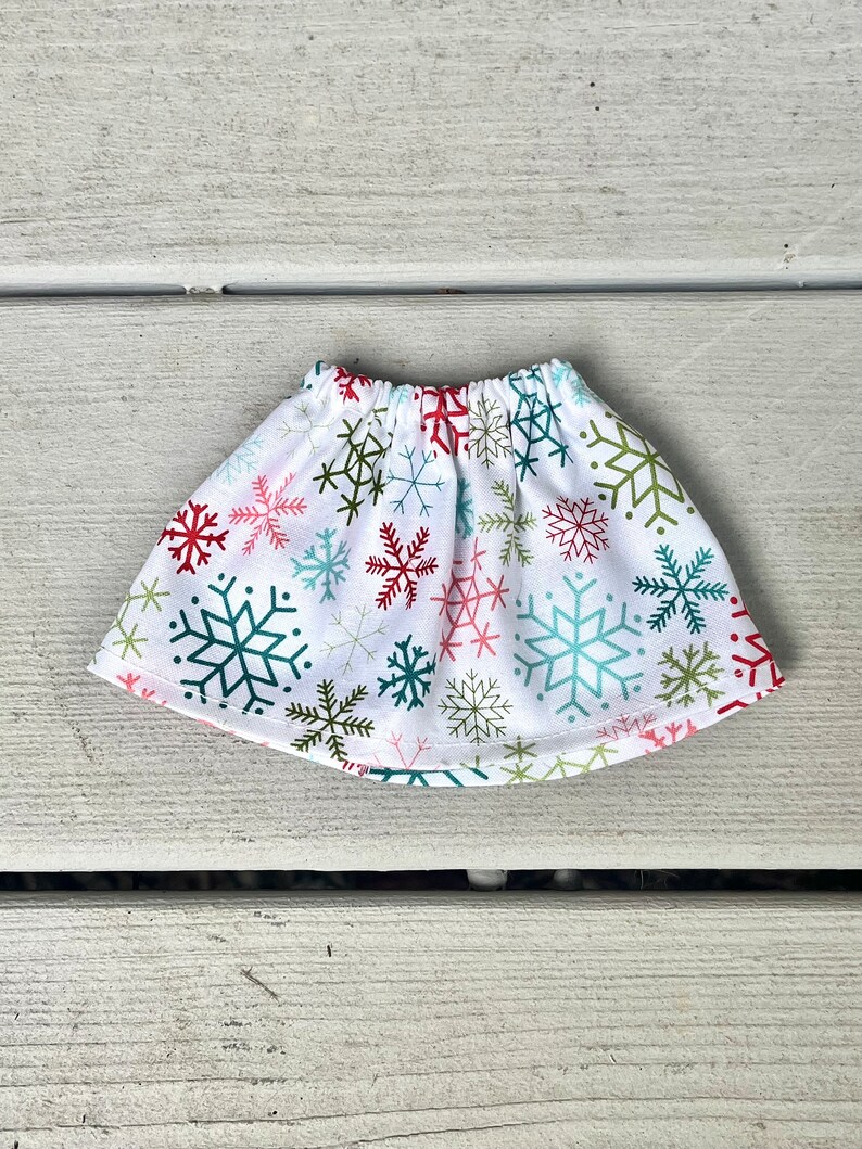Puede incluir: Una falda blanca con un patr&oacute;n de copo de nieve en rojo, verde y azul. La falda tiene una cintura el&aacute;stica.