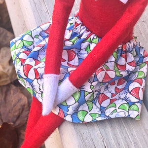 Puede incluir: Mu&ntilde;eca de duende con una camisa roja de manga larga y una falda con estampado de bastones de caramelo azul y blanco.