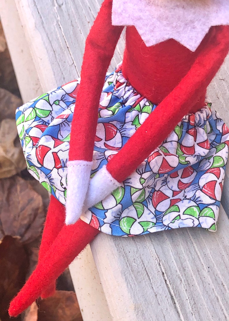 Puede incluir: Mu&ntilde;eca de duende con una camisa roja de manga larga y una falda con estampado de bastones de caramelo azul y blanco.