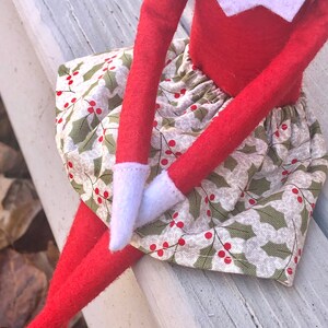 Puede incluir: Una mu&ntilde;eca de elfo roja con una camisa de cuello blanco y una falda verde y blanca con bayas de acebo rojas. La mu&ntilde;eca est&aacute; sentada en un banco de madera blanco.