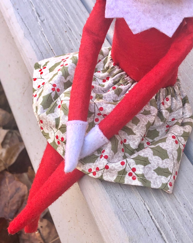Puede incluir: Una mu&ntilde;eca de elfo roja con una camisa de cuello blanco y una falda verde y blanca con bayas de acebo rojas. La mu&ntilde;eca est&aacute; sentada en un banco de madera blanco.