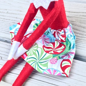 Puede incluir: Una mu&ntilde;eca de elfo roja con una camisa de cuello blanco y una falda de estampado de bast&oacute;n de caramelo de colores. La falda tiene un fondo blanco con bastones de caramelo rojos, verdes, azules y rosas.