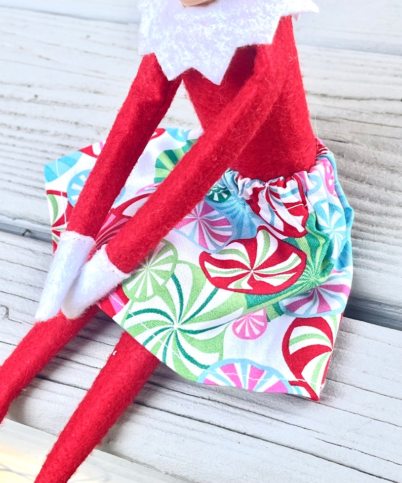 Puede incluir: Una mu&ntilde;eca de elfo roja con una camisa de cuello blanco y una falda de estampado de bast&oacute;n de caramelo de colores. La falda tiene un fondo blanco con bastones de caramelo rojos, verdes, azules y rosas.