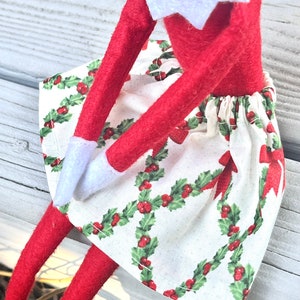 Puede incluir: Una mu&ntilde;eca de elfo roja con un cuello blanco y una falda con estampado de acebo blanco y rojo.