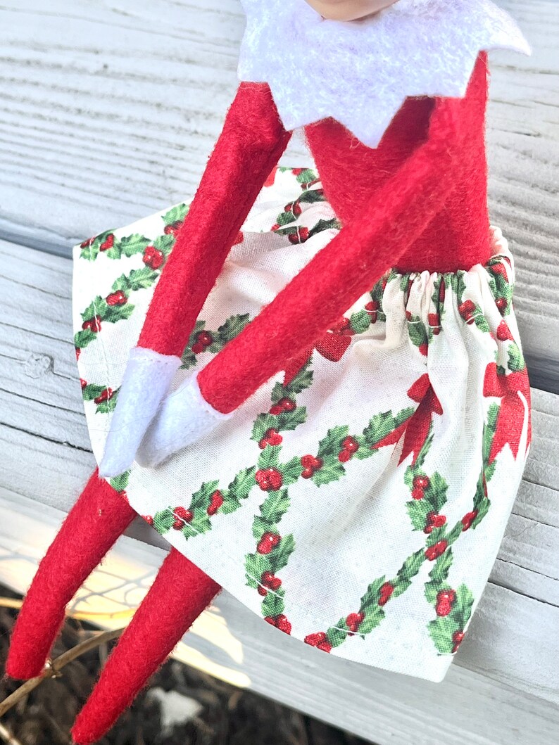 Puede incluir: Una mu&ntilde;eca de elfo roja con un cuello blanco y una falda con estampado de acebo blanco y rojo.
