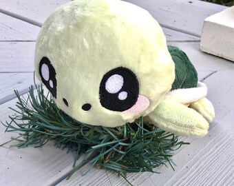 May include: A plush green turtle with large, black eyes and a pink blush. The turtle is lying on a bed of green pine needles.