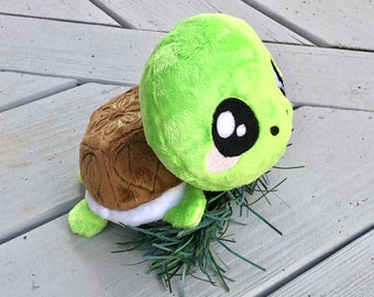 May include: A plush turtle toy with a green head and limbs, a brown shell with embroidered details, and large black and white eyes. The toy is resting on a bed of green faux grass, creating a whimsical scene.