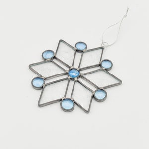 May include: A stained glass snowflake ornament with a silver-colored metal frame. The snowflake features clear, textured glass panes and light blue glass beads at the points and center. A silver string is attached for hanging.
