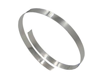 May include: A thin, flexible, silver-colored metal strip formed into an open, overlapping circular coil. The material has a smooth, reflective surface with a metallic sheen, showing a continuous band that curves to create a simple, circular shape.
