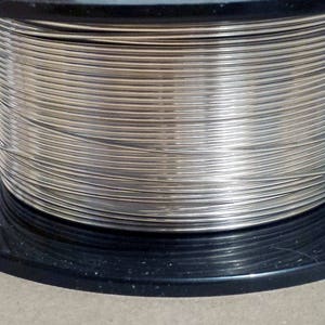 May include: A spool of silver-colored wire is tightly wound on a black plastic reel. The wire appears to be thin and flexible, suitable for crafting or jewelry making. The reel sits on a neutral-toned surface.