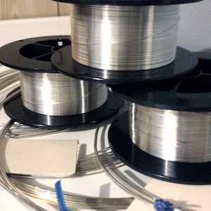 May include: Several spools of silver-colored wire are stacked on top of each other. The wire is wound around black plastic spools. Some loose wire is visible in the foreground. A small, folded piece of cardboard and a blue ribbon are also present.