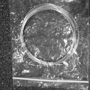 May include: A bundle of silver-colored wire, coiled and secured with tape, inside a clear plastic bag. The wire is thin and appears to be made of metal. The bag is sealed with a zipper closure. The background is a dark, textured surface.