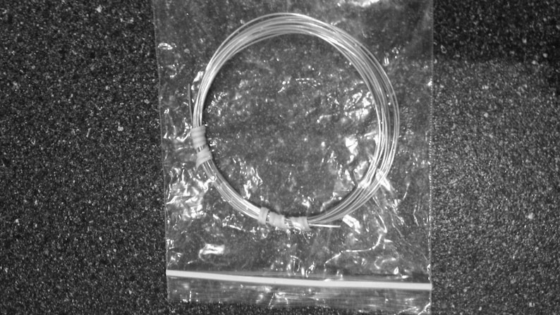 May include: A bundle of silver-colored wire, coiled and secured with tape, inside a clear plastic bag. The wire is thin and appears to be made of metal. The bag is sealed with a zipper closure. The background is a dark, textured surface.
