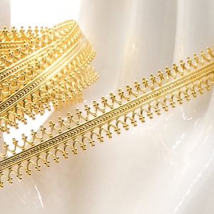 May include: Close-up of a gold-colored decorative trim. The trim features a repeating pattern of small arches and beaded details along its edges. The trim is rolled up, showcasing its intricate design and metallic sheen, suitable for crafts and embellishments.