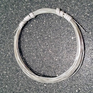 May include: A coil of silver-colored wire, tightly wound into a circular shape. The wire is bundled and secured with white tape at two points. The image is taken against a dark, textured background, highlighting the metallic sheen of the wire.