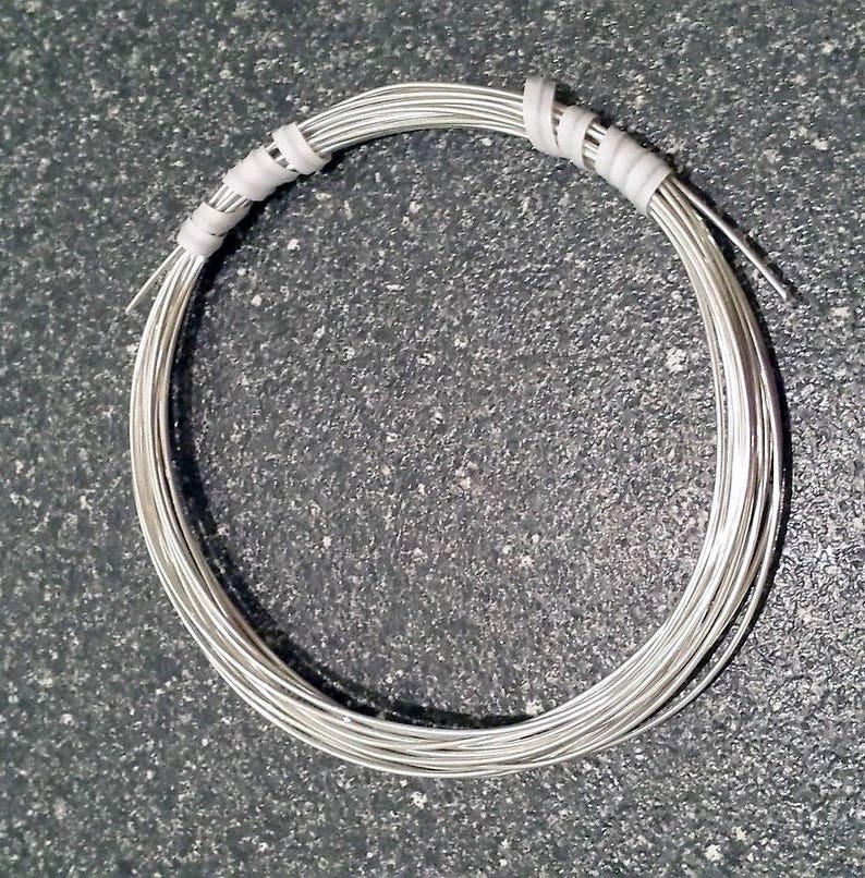 May include: A coil of silver-colored wire, tightly wound into a circular shape. The wire is bundled and secured with white tape at two points. The image is taken against a dark, textured background, highlighting the metallic sheen of the wire.