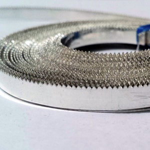 May include: A coiled roll of silver-colored metal tape with a serrated edge. The tape is tightly wound, revealing the textured surface and sharp teeth along the outer rim. The background is a soft white.