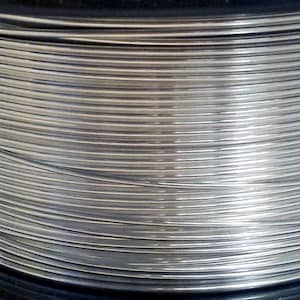 May include: Close-up of a spool of silver-colored wire. The wire is tightly wound, creating a pattern of parallel lines. The metallic sheen reflects light, highlighting the texture and form of the wire. The spool is set against a dark background.