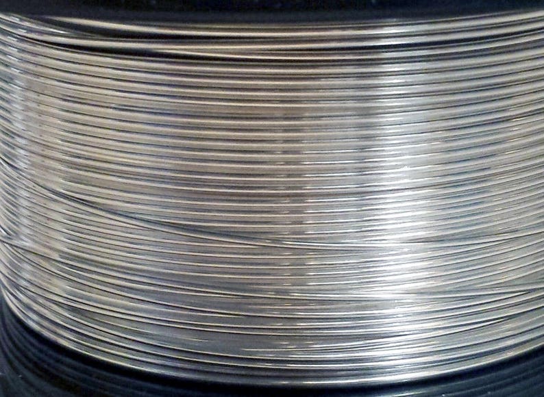 May include: Close-up of a spool of silver-colored wire. The wire is tightly wound, creating a pattern of parallel lines. The metallic sheen reflects light, highlighting the texture and form of the wire. The spool is set against a dark background.