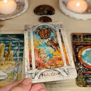 May include: A collection of tarot cards with colorful illustrations, including a card with a turtle and the word "PROTECTION". Two lit candles in seashell holders and other decorative elements are in the background.