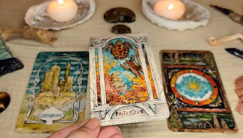 May include: A collection of tarot cards with colorful illustrations, including a card with a turtle and the word "PROTECTION". Two lit candles in seashell holders and other decorative elements are in the background.