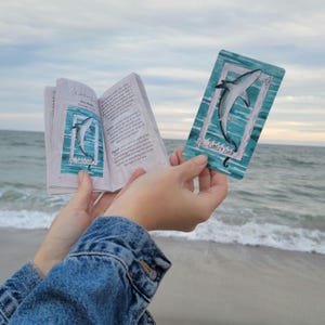 May include: An open book and a card featuring a shark illustration. The book's pages display text and a shark image within a rectangular frame. The card shows a shark in a similar frame, with the words "Comfort Zone" at the bottom. The background is a beach scene.