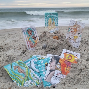 May include: A collection of tarot cards with colorful illustrations, including a seahorse, turtle, and flamingo, arranged on a sandy beach with the ocean in the background. Some cards have text that reads "Hold On" and "Listen".