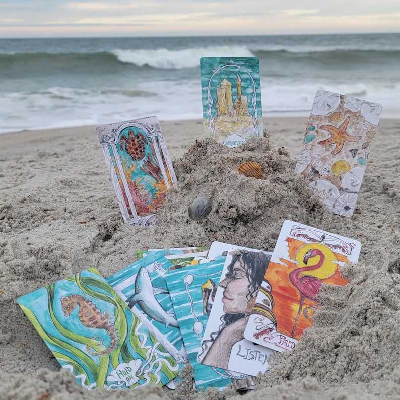 May include: A collection of tarot cards with colorful illustrations, including a seahorse, turtle, and flamingo, arranged on a sandy beach with the ocean in the background. Some cards have text that reads "Hold On" and "Listen".