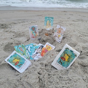 May include: A collection of oracle cards and a box set on a sandy beach. The cards feature colorful illustrations of sea creatures, architectural structures, and abstract designs. The box set has the text "Floridian Mitos Oracle Deck" with a whimsical illustration.