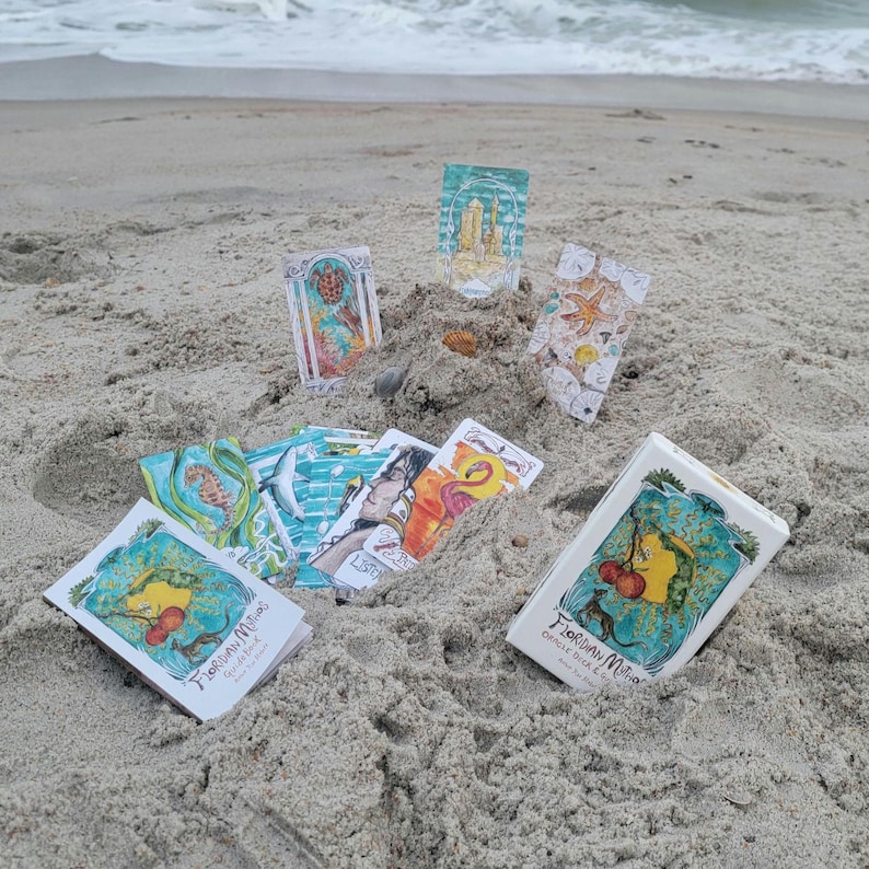 May include: A collection of oracle cards and a box set on a sandy beach. The cards feature colorful illustrations of sea creatures, architectural structures, and abstract designs. The box set has the text "Floridian Mitos Oracle Deck" with a whimsical illustration.