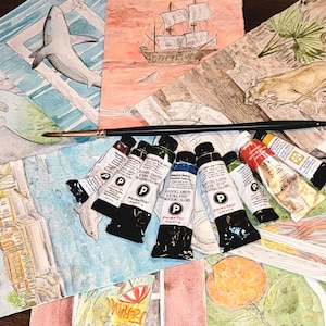 May include: A collection of watercolor paintings featuring marine life, a sailboat, and a lighthouse. A paintbrush and tubes of paint are arranged on top of the paintings. The paintings are in various sizes and colors, with a focus on aquatic themes.